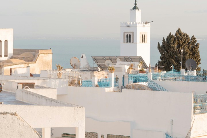 White buildings with blue accents in a coastal town, featuring a tall tower and distant sea view.