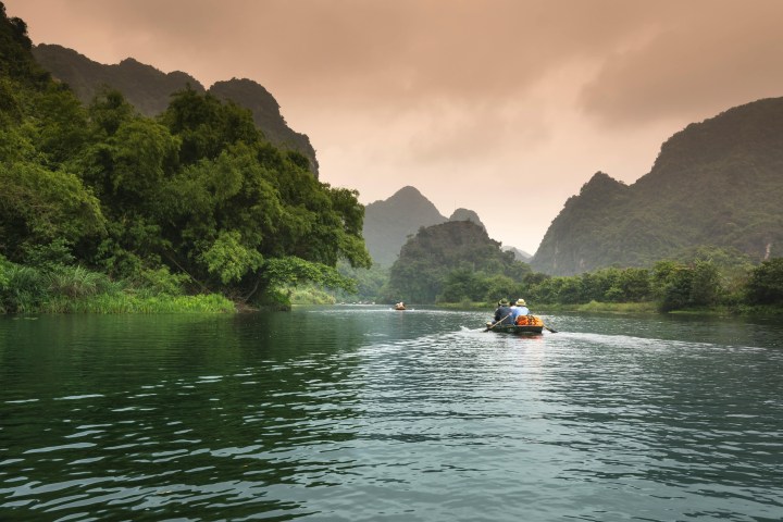 People rowing boats on a calm river with lush greenery and mountains under a cloudy sky.