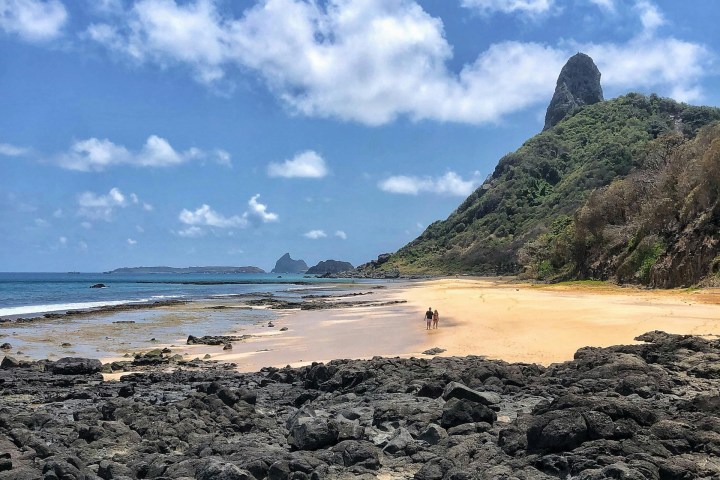 Rocky beach with hills, clear sky, and distant people walking on sand under scattered clouds.