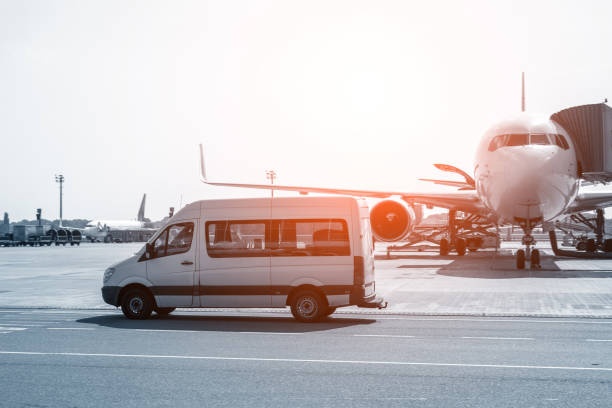 White van driving by a parked airplane at an airport under bright sunlight.