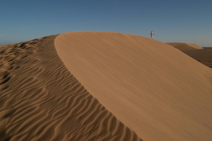 Person in white clothing stands on a large sand dune under a clear blue sky.
