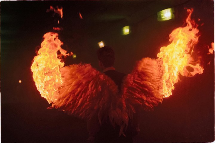 Person with wings on fire in a dimly lit setting, facing away.