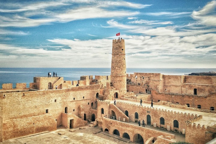 Ancient stone fortress with a central tower, surrounded by people and sea under a partly cloudy sky.