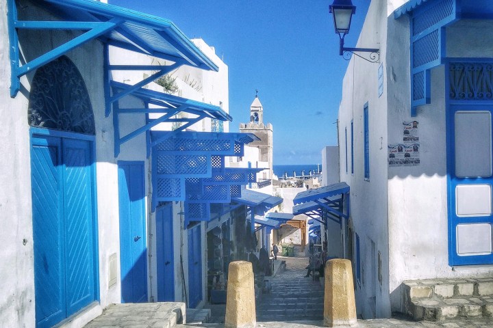 Narrow cobblestone street with white buildings and blue accents under a clear blue sky.
