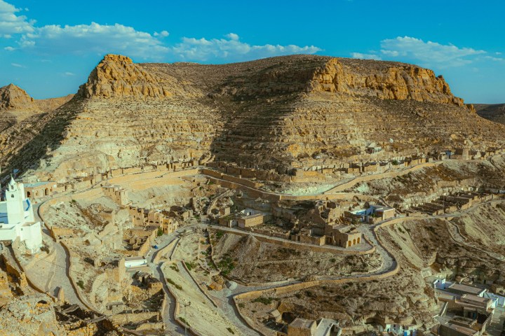 Aerial view of ancient mountain village with white buildings and rugged terrain under blue sky.